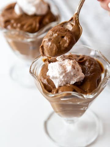A hand holding a silver spoon with chocolate pudding and 2 glass dishes of chocolate pudding with whipped topping.