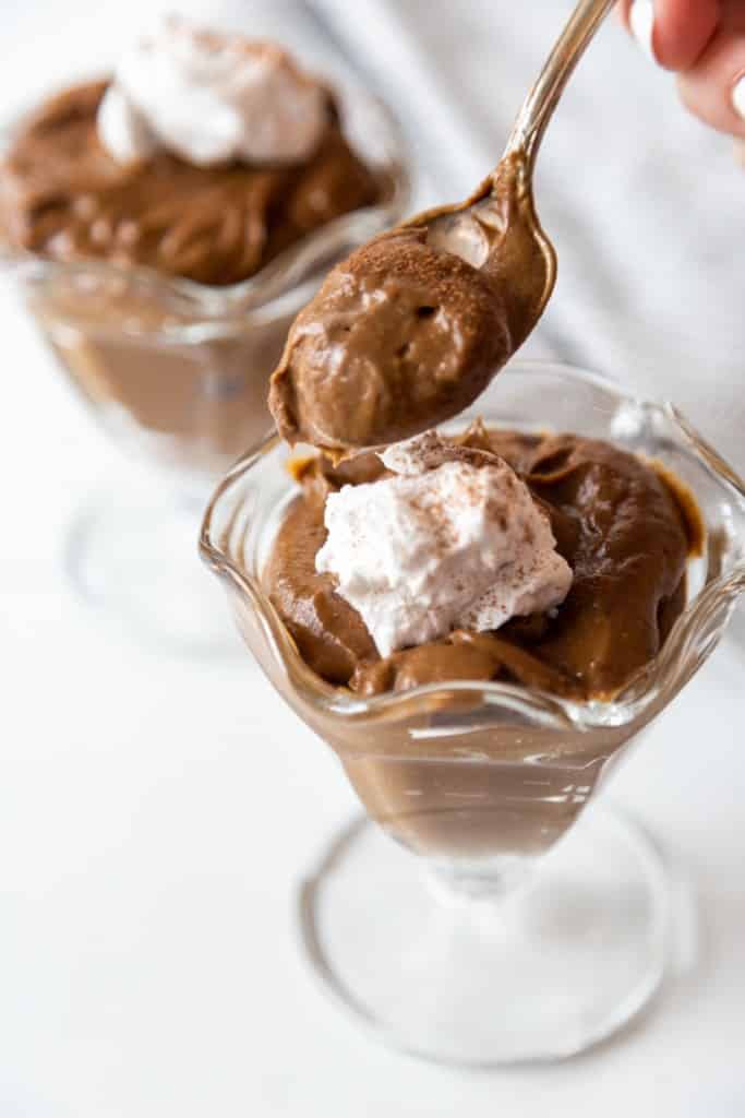 A hand holding a silver spoon with chocolate pudding and 2 glass dishes of chocolate pudding with whipped topping.