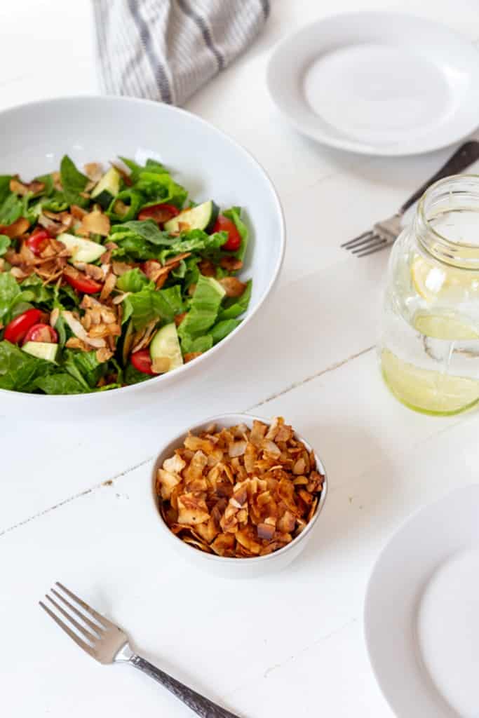 A salad in a white bowl with a small white bowl of coconut bacon and white plates and silver forks next to the bowls.