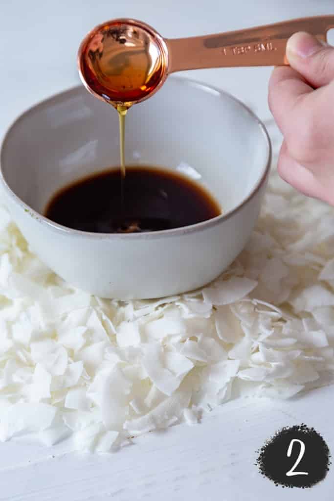 A hand pouring maple syrup into a bowl of liquid smoke with a copper measuring spoon.