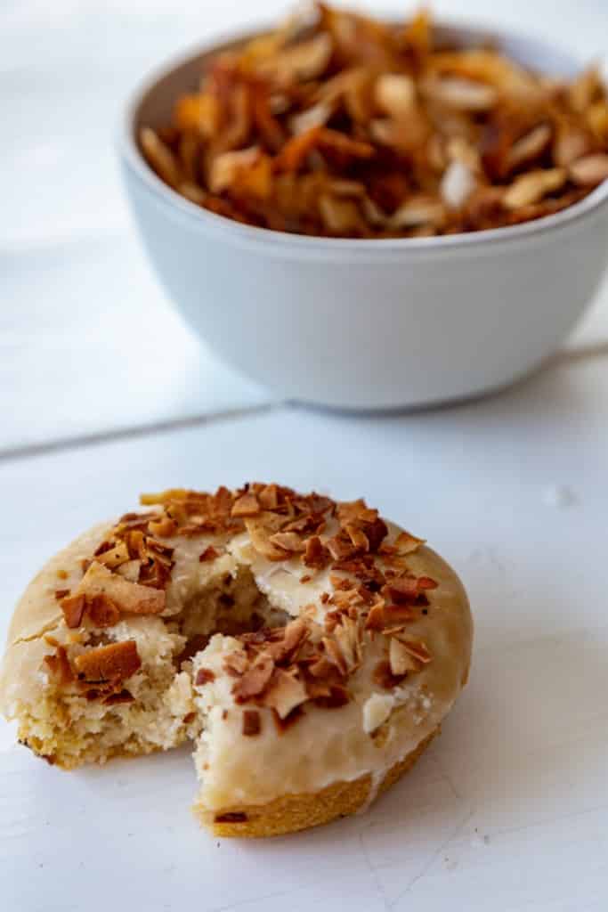 A partially eaten doughnut with coconut bacon on top and a white bowl with the bacon.