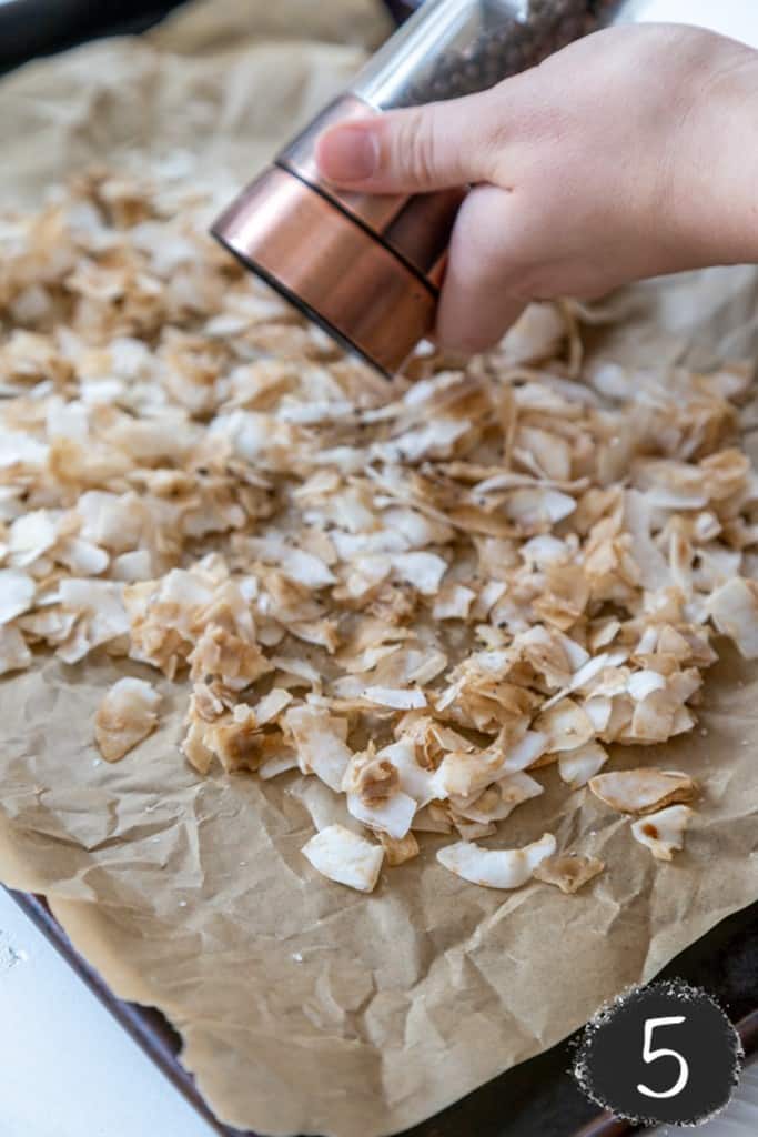 A baking sheet lined with parchment paper and coconut flakes that are coated in a brown liquid spread on the pan. A hand is grinding pepper over the coconut.