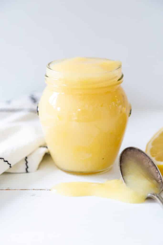 A jar of vegan lemon curd and a spoon dripping curd on a white table is next to the jar.