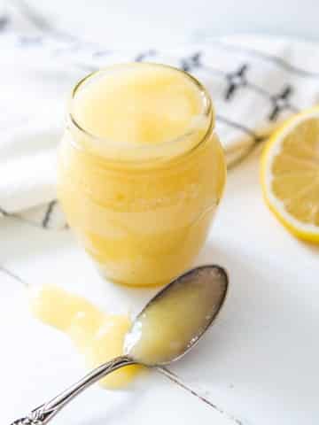 A glass jar of lemon curd with a silver spoon full of the curd sitting on a white wood surface.