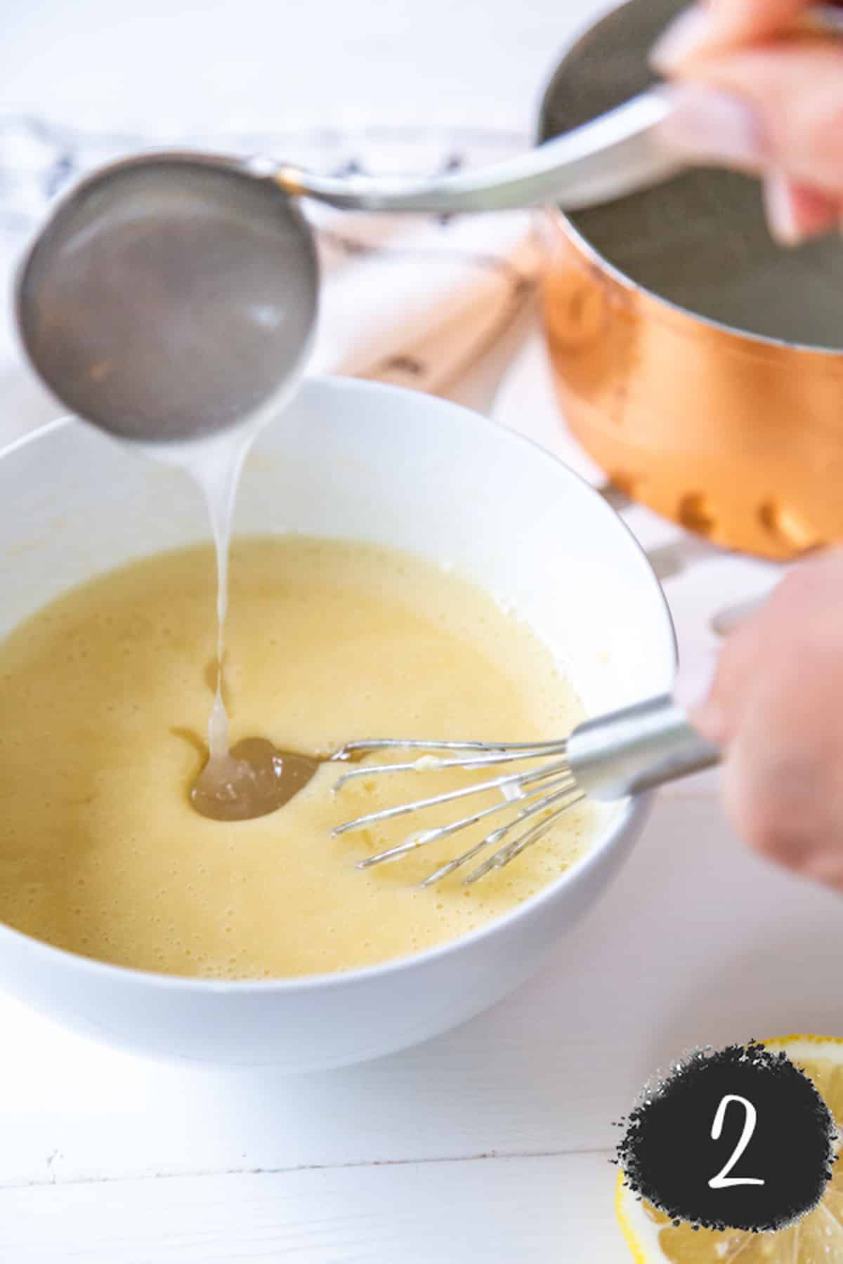 A white bowl with and egg mixture and a hand pouring sugar water into the bowl and whisking the mixture.