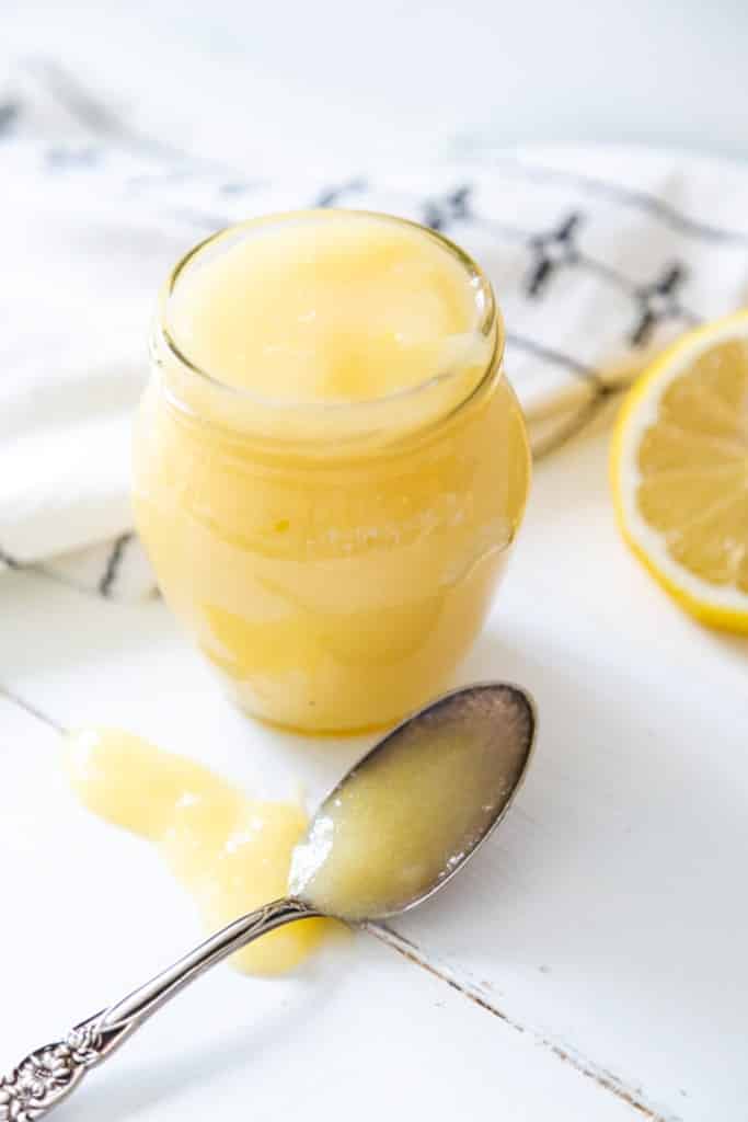 A glass jar of lemon curd with a silver spoon full of the curd sitting on a white wood surface.