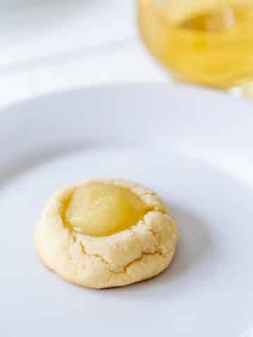 A lemon filled thumbprint cookie on a white plate with a glass of wine next to the plate.