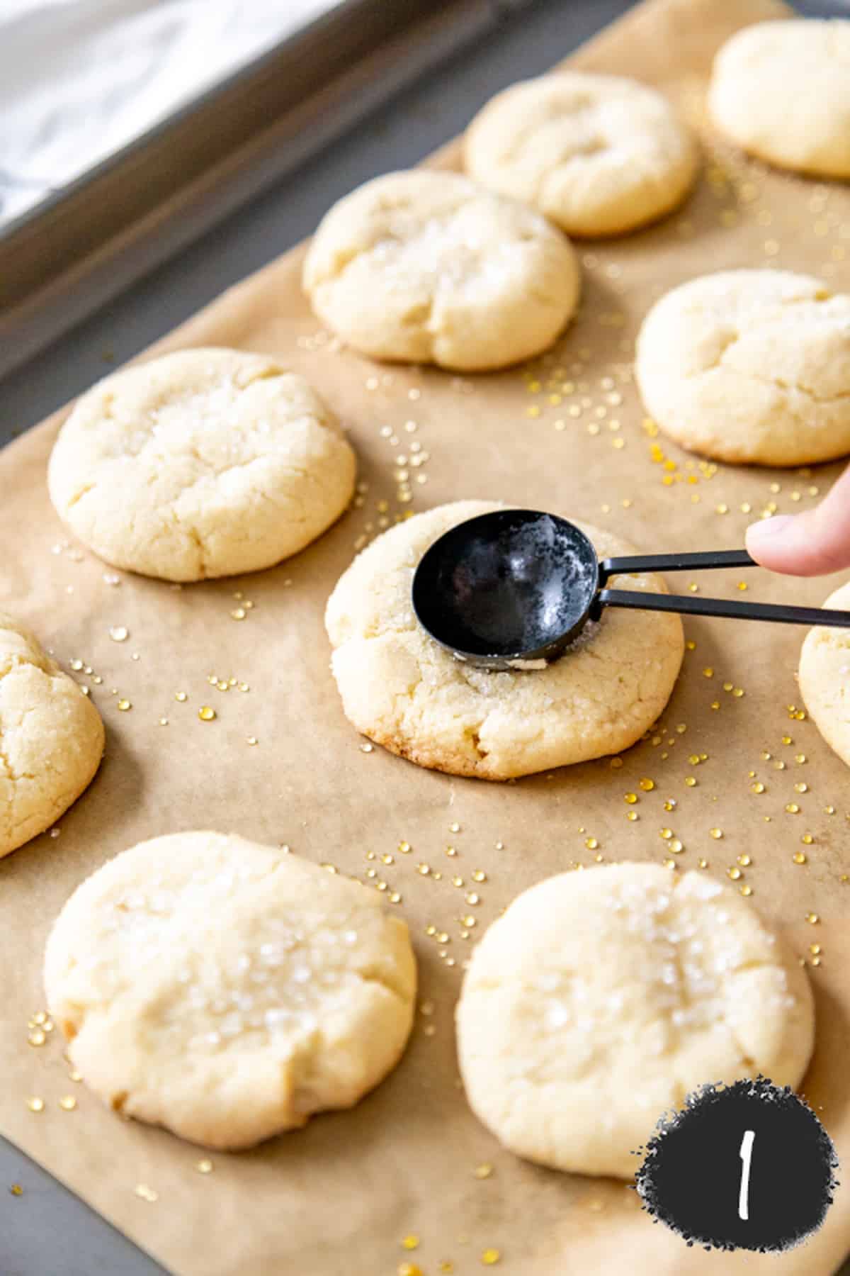 A baking sheet with sugar cookies and a hand pressing a black measuring spoon into the center of the cookie to make and indentation.