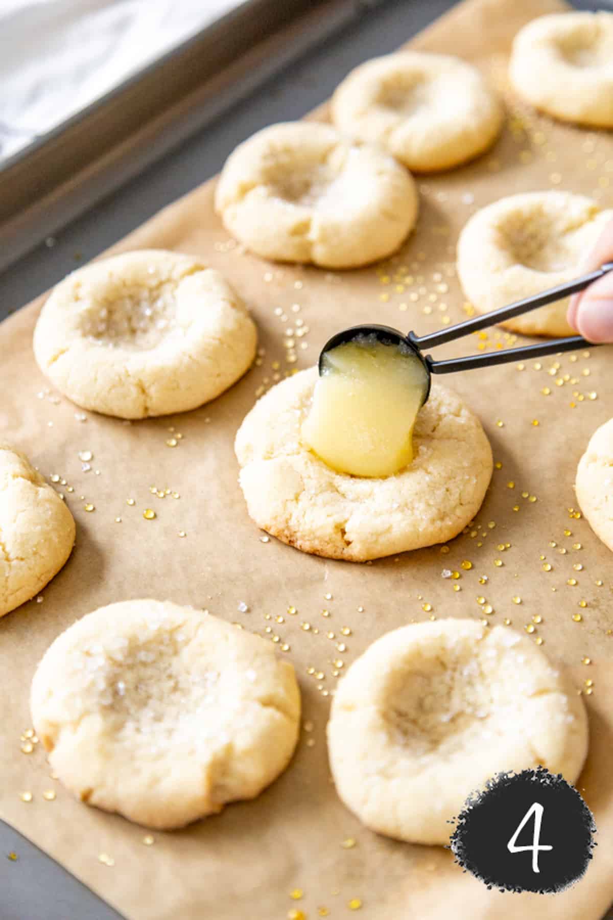 A cookie sheet with sugar cookies with indentations in the centers and a hand dropping lemon curd in the center of one of the cookies with a black measuring spoon.
