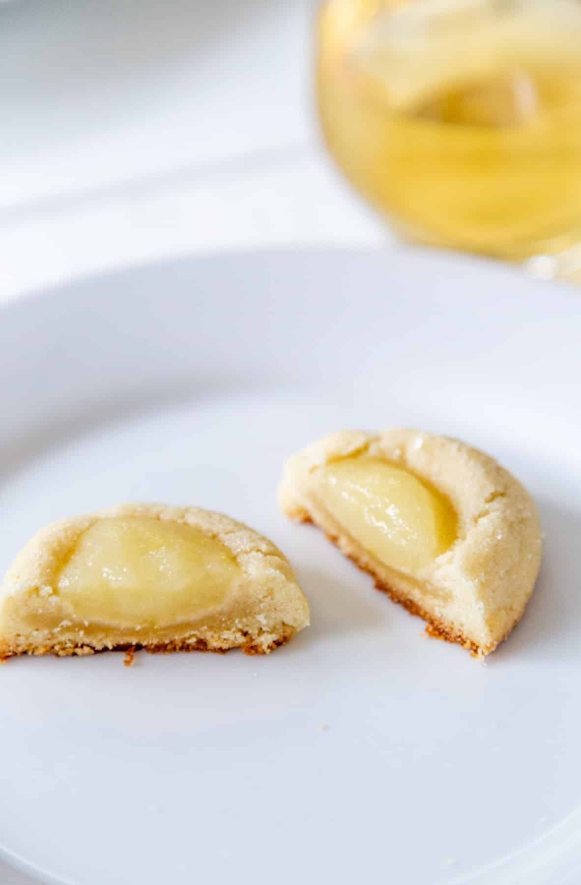 A lemon thumbprint cookie broken in half on a white plate with a glass of wine in the background.