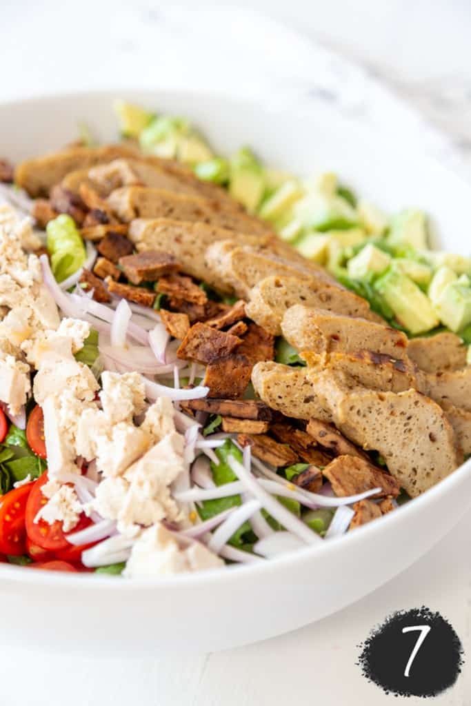 A white salad bowl with lettuce, bacon bites, slivered onions and halved cherry tomatoes, sliced vegan chicken, avocado, and vegan cheese.