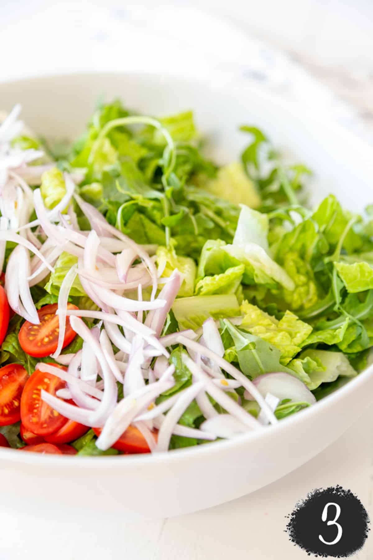 A white salad bowl with lettuce, tomatoes, and slivered onions.