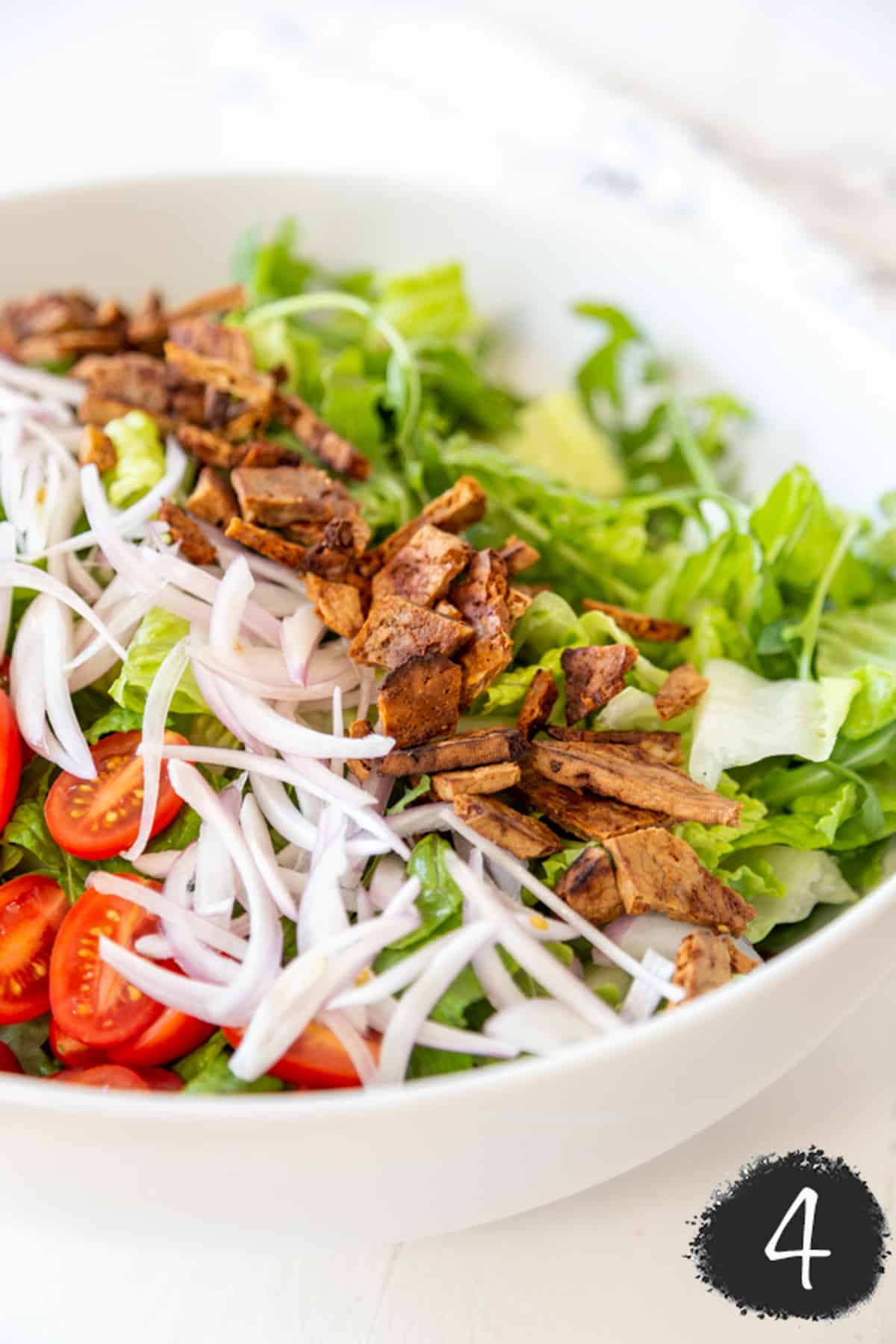 A white salad bowl with lettuce, bacon bites, slivered onions and halved cherry tomatoes.