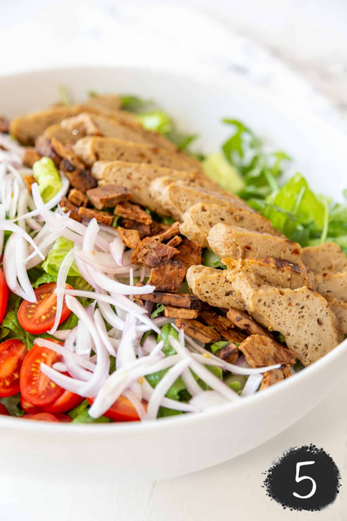 A white salad bowl with lettuce, bacon bites, slivered onions and halved cherry tomatoes, and sliced vegan chicken.