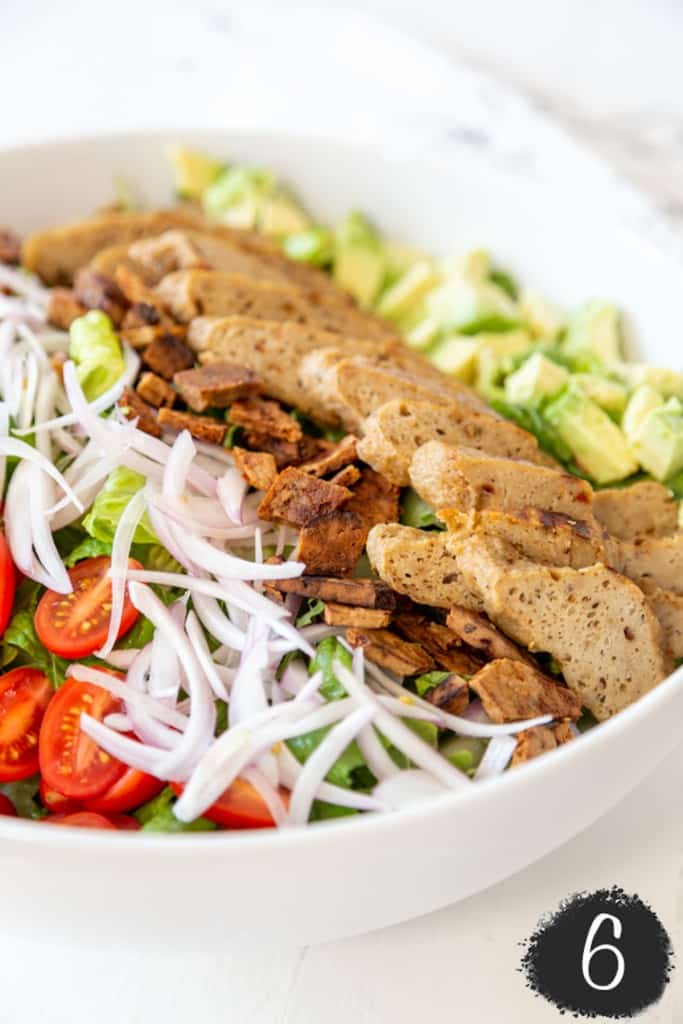 A white salad bowl with lettuce, bacon bites, slivered onions and halved cherry tomatoes, sliced vegan chicken, and cubed avocado.