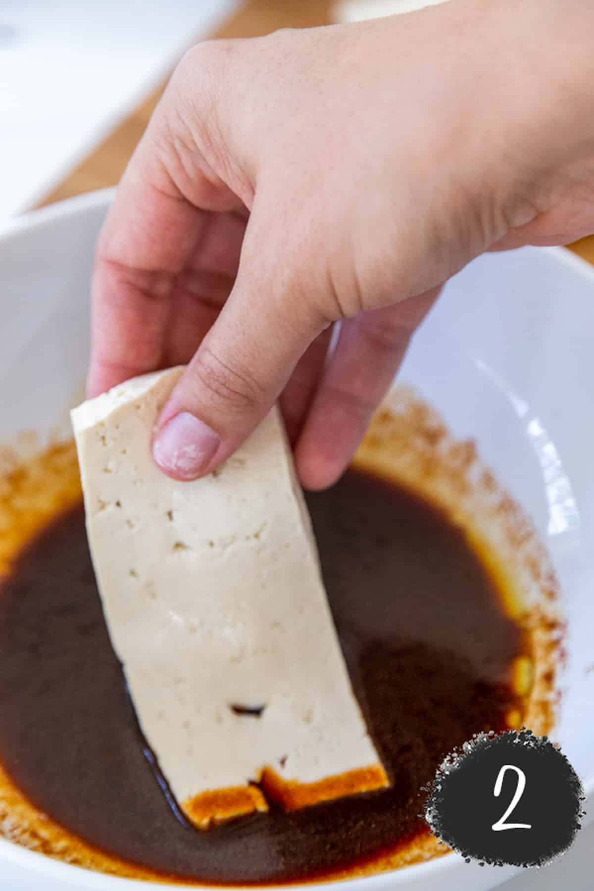 A hand dipping a slice of tofu into a white bowl filled with brown sauce.