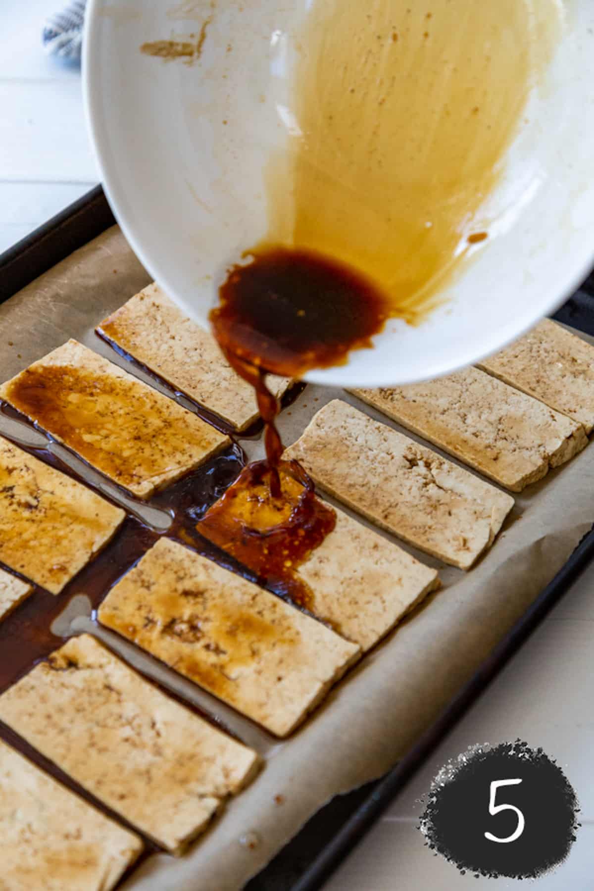 A white bowl with soy sauce being poured over sliced tofu on a baking sheet.
