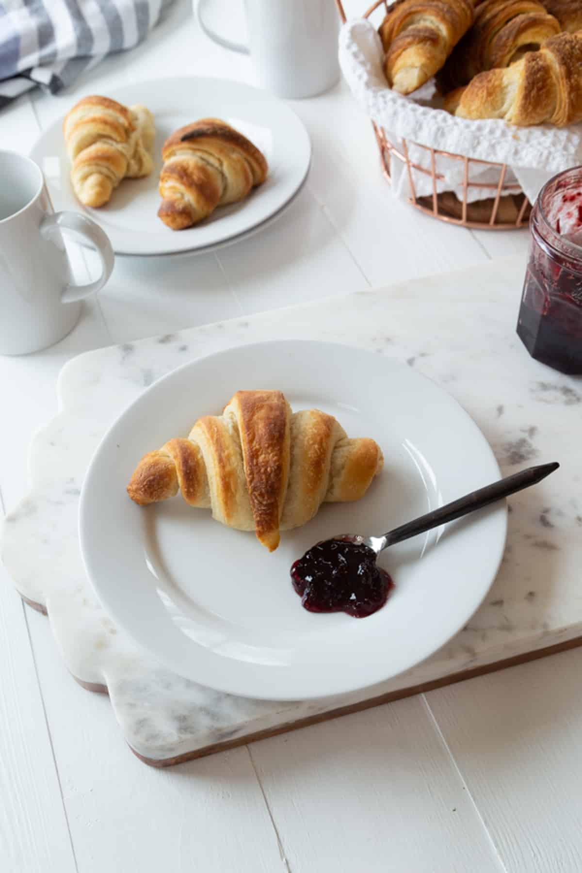 A plate with a croissant and jam and a basket of croissants in the background.