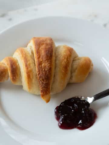 A croissant on a white plate with a spoon of preserves.