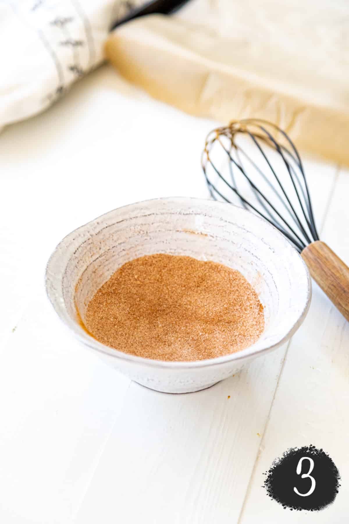 A white bowl with cinnamon sugar and a whisk.