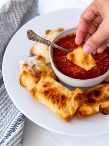 A hand dipping a pizza roll into a bowl of marinara sauce.