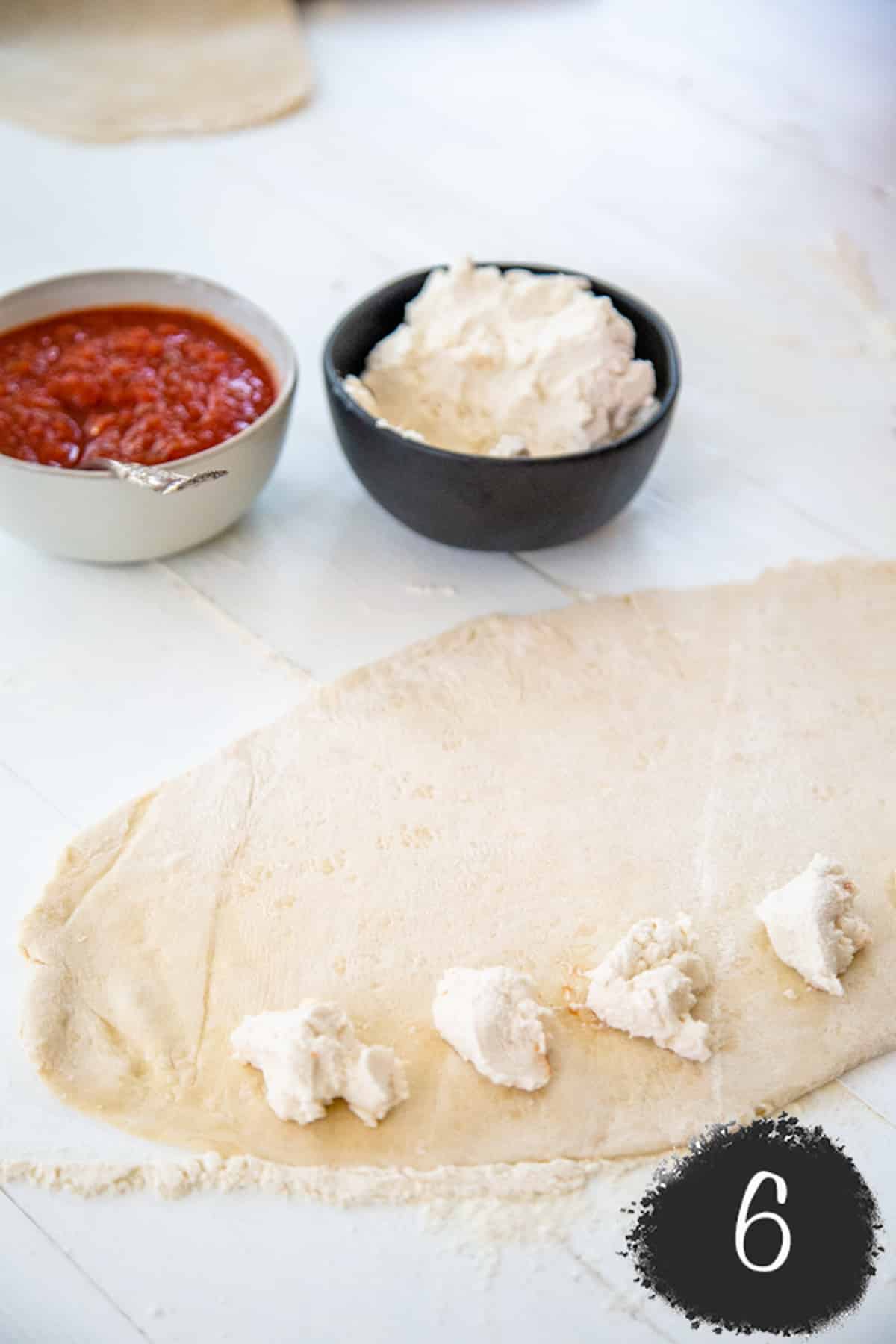 Dollops of cheese spread evenly on rolled out pizza dough.