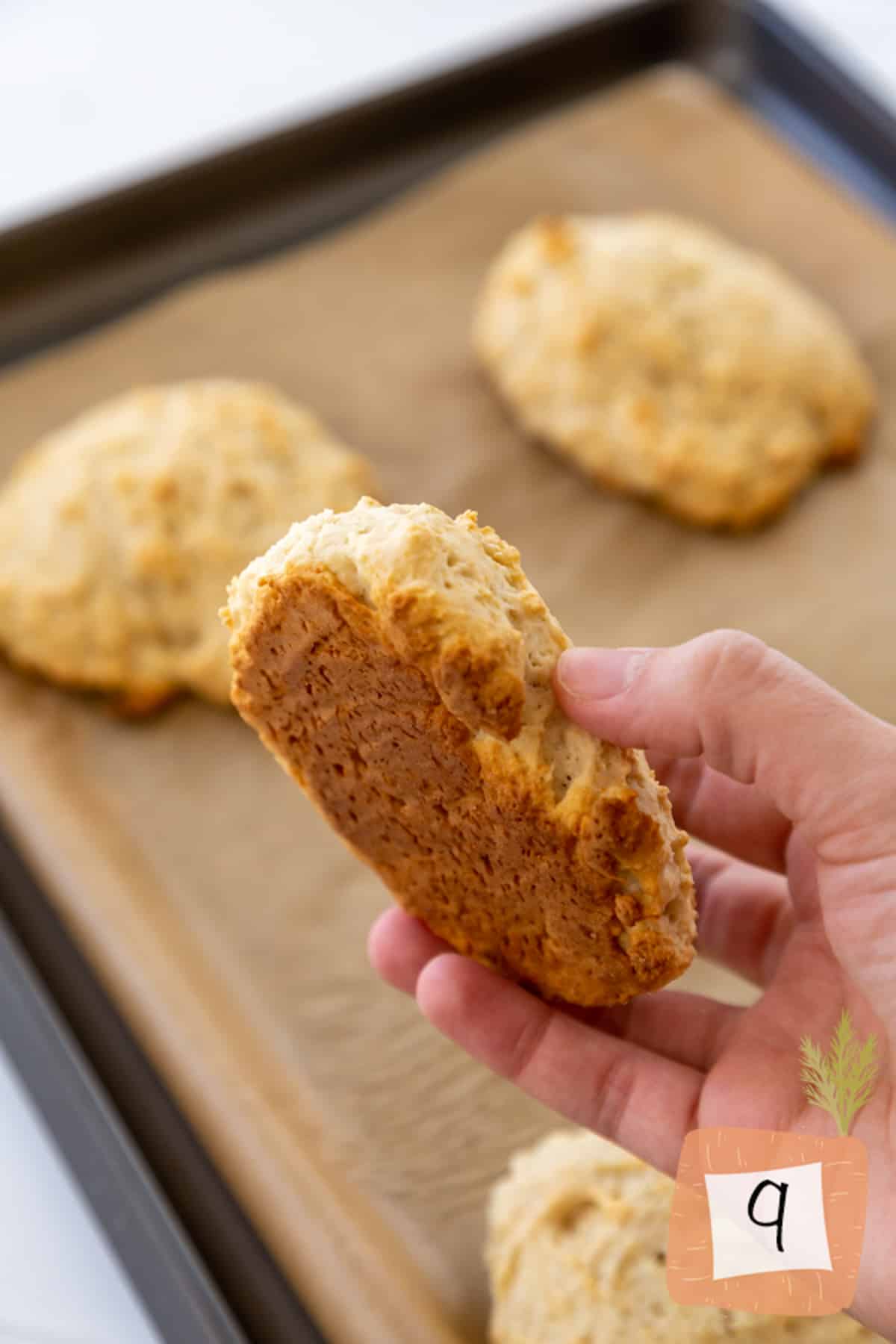 A hand holding a biscuit with a golden brown bottom over a baking sheet.