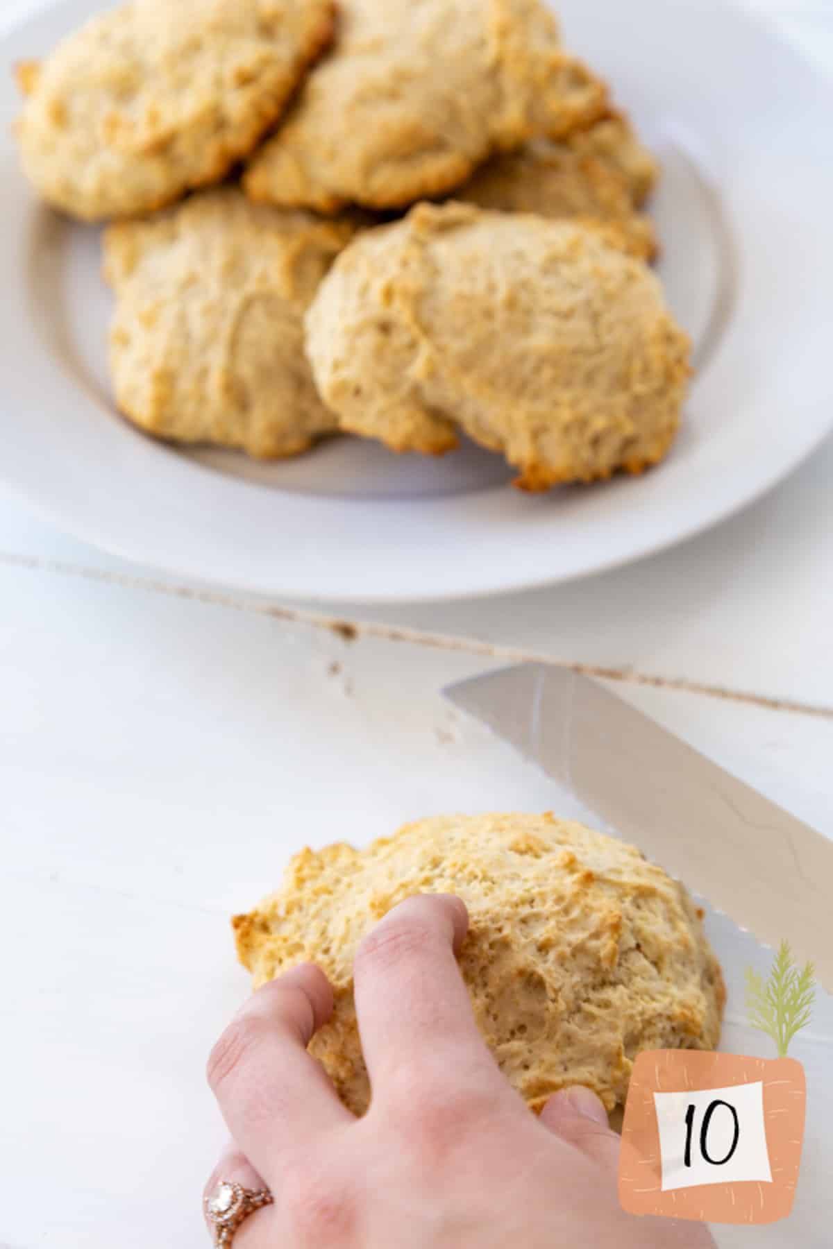 A hand holding a drop biscuit with a white plate of biscuits in the background.