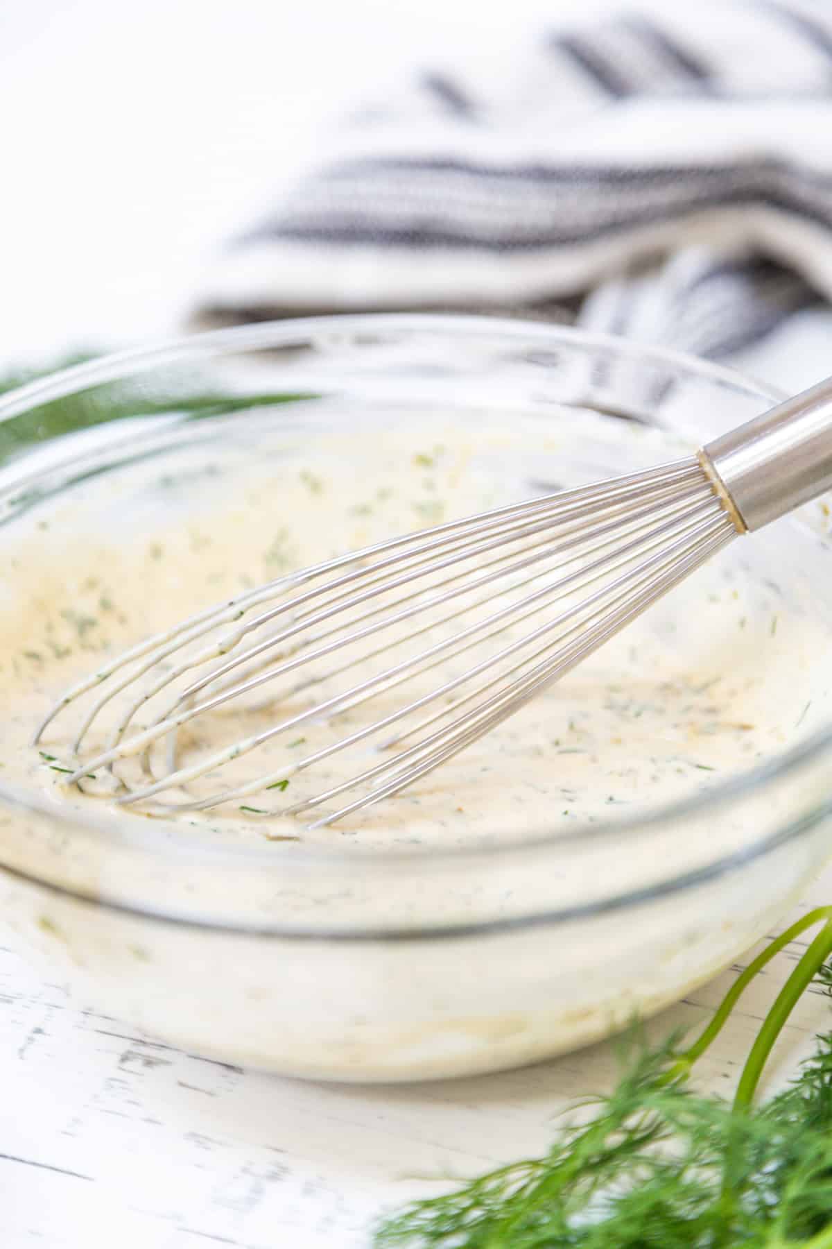 A clear glass bowl of tartar sauce with a whisk.
