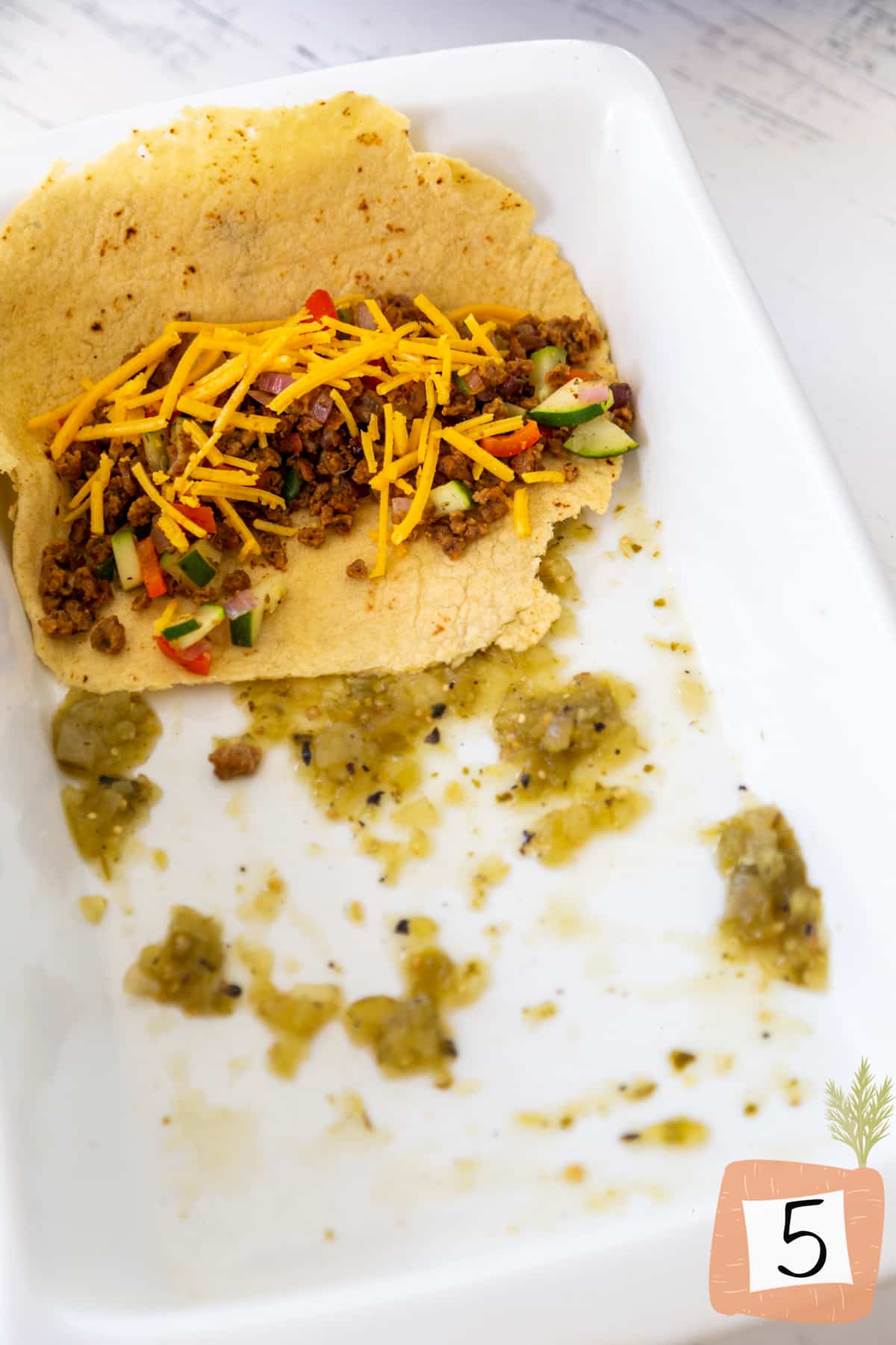 A white baking dish with green salsa and a corn tortilla filled with vegetables, vegan chorizo, and vegan cheese.